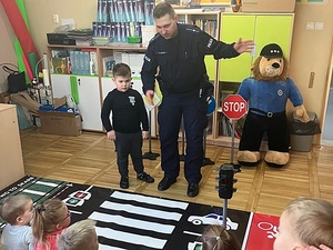 sala w przedszkolu, policjant pokazuje dziecku matę edukacyjną