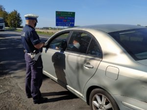 funkcjonariusz Policji stoi przy samochodzie koloru srebrnego, wewnątrz za kierownicą siedzi mężczyzna