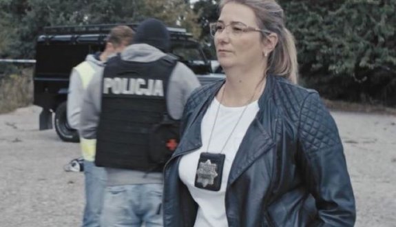 Kobieta z zawieszoną na szyi odznaką policyjną, w tle mężczyzna w czarnej kamizelce z napisem POLICJ