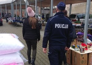 policjant na placu targowym, przed nim idzie kobieta, obok widać stragany