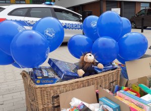 niebieskie balony, maskotka, oraz zabawki w kartonach, za nimi w tle radiowóz