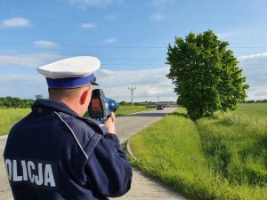 policjant stoi przy drodze, w dłoniach trzyma miernik prędkości , w tle droga